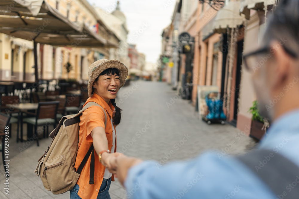 Fototapeta premium Tourist woman leading her partner by hand while walking through a city street lined with cafes and shops