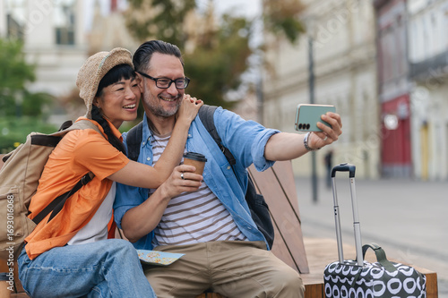 Happy middle-aged tourists sitting on a bench with luggage, taking a cheerful selfie while exploring a historic European city on vacation