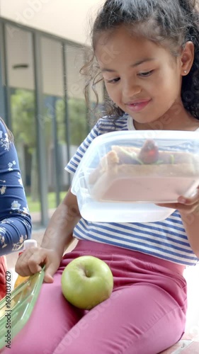 Vertical video: Diverse girl friends preparing lunchbox on patio, arranging sandwich, placing apple