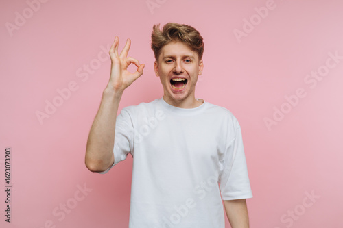 Young man smiles while making an OK gesture against a pastel pink background in a cheerful setting