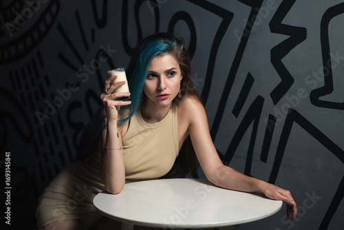 A stylish young woman with blue-highlighted hair poses at a modern café table, holding a drink against an artistic black-and-white background