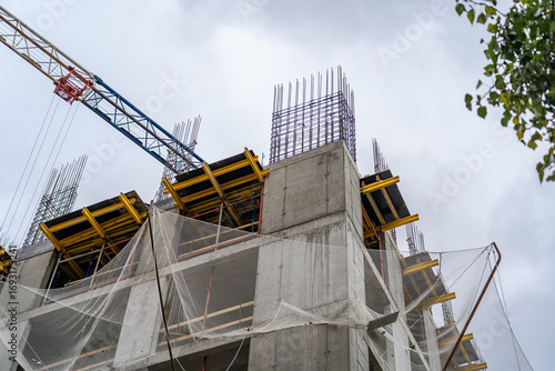 Dynamic low-angle perspective of multi-story building under construction, featuring raw concrete, exposed rebar, temporary yellow formwork, protective safety nets, and prominent tower crane against ov