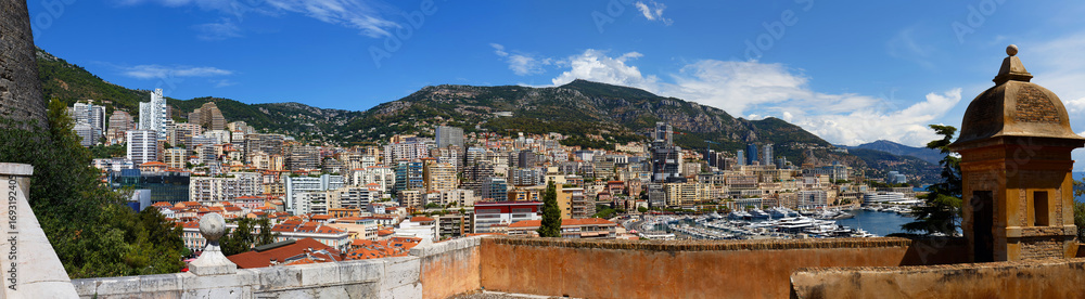 Naklejka premium Elevated view of Monaco cityscape with red rooftops, layered high-rises, and lush green hills. Scenic urban landscape of the Principality, blending modern architecture with Mediterranean charm.