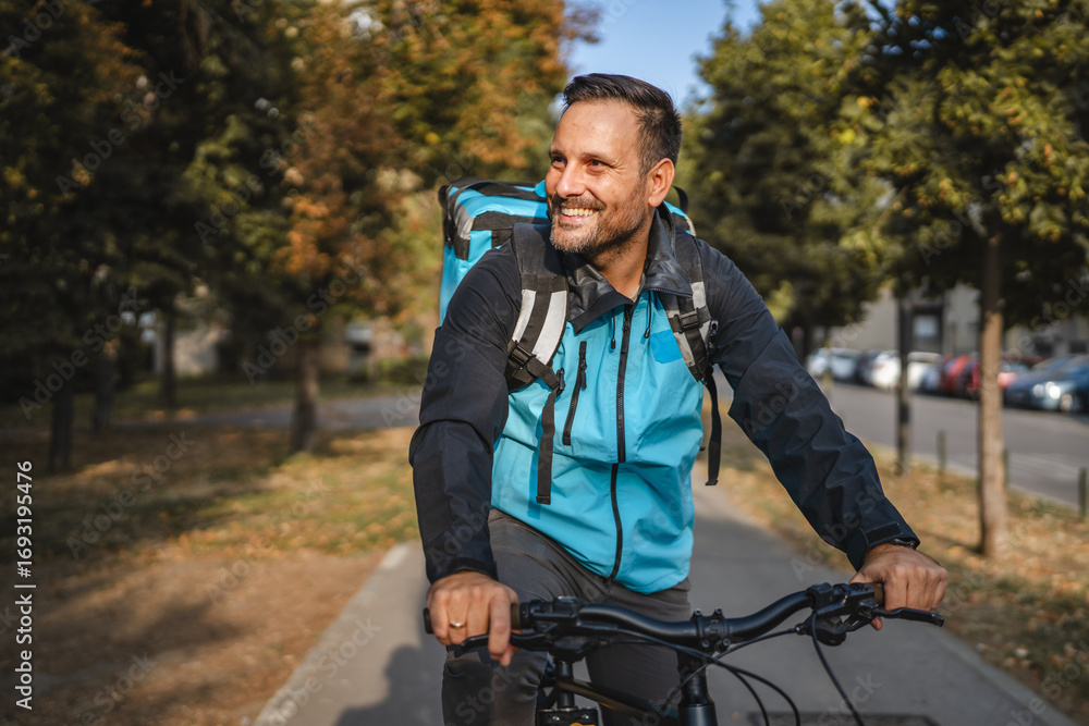Fototapeta premium Delivery man riding bicycle smiling carrying insulated backpack in city park