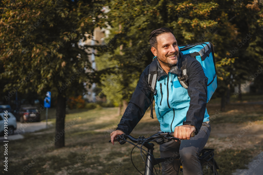 Fototapeta premium Delivery man riding bicycle smiling carrying insulated backpack in city park