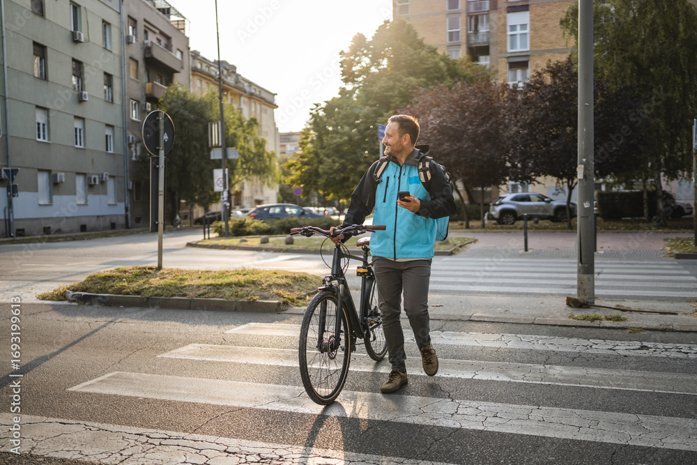 Fototapeta premium Delivery man crossing the street with bicycle and smart phone at sunset