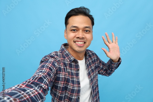 Happy smiling young Asian man, wearing casual clothes, POV selfie on mobile phone, waving, greeting someone, isolated on blue background.