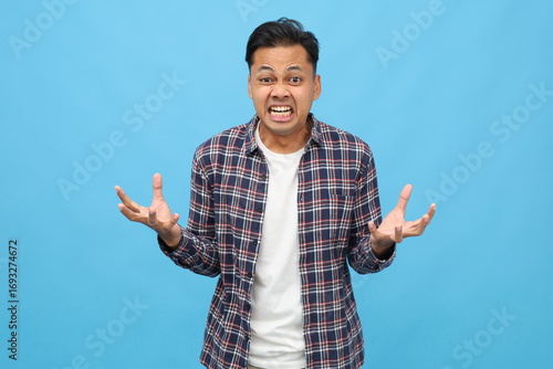 An annoyed and angry man raises his hands and grits his teeth in anger, staring at the camera in annoyance, about to kill someone, standing on a blue background.