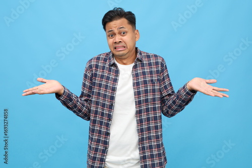 Fotografie Sad young asian man wearing casual clothes, spreading his arms, shrugging his shoulders, looking confused, clueless, isolated on blue background