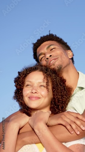 Vertical video: Diverse couple hugging under clear sky while expressing affection and gazing upward