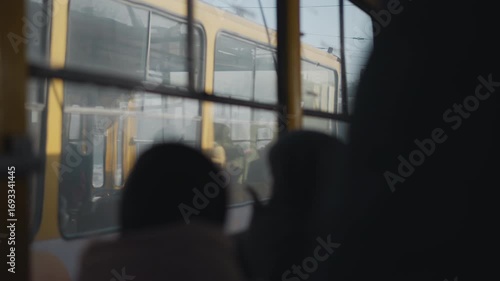 Inside moving bus, passengers in winter coats watch opposite tram glide past outside, view through windows with soft blur on glass, muted city light, motion on snowy street during urban commute