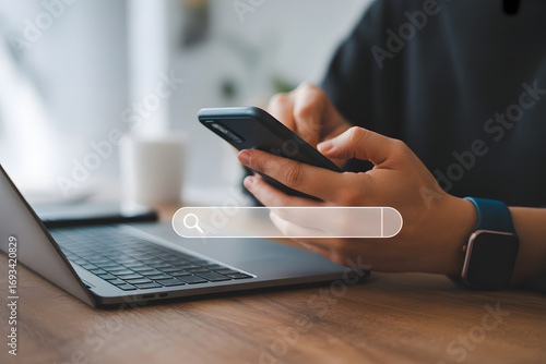 Digital Interaction: A person focused on a search bar on a phone interacting with laptop to perform an online query in a well-lit, focused workspace