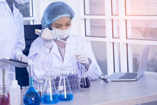 Focused female scientist in a white lab coat, wearing a hairnet, mask, and safety glasses, meticulously works with test tubes and beakers in a sunlit laboratory.