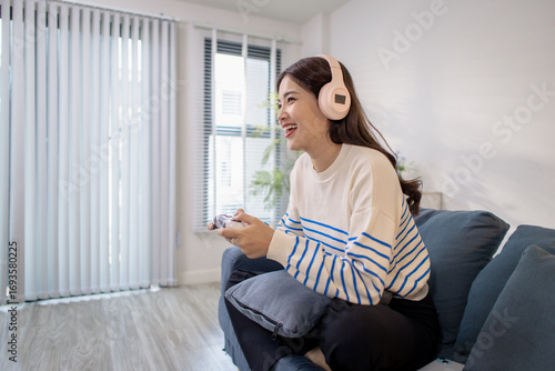 A young woman sits on a sofa wearing headphones while holding a game controller and playing a video game on her laptop. Her cheerful expression shows enjoyment and excitement. Free Download A young woman sits on a sofa wearing headphones while holding a game controller and playing a video game on her laptop. Her cheerful expression shows enjoyment and excitement. - GFXLoad
