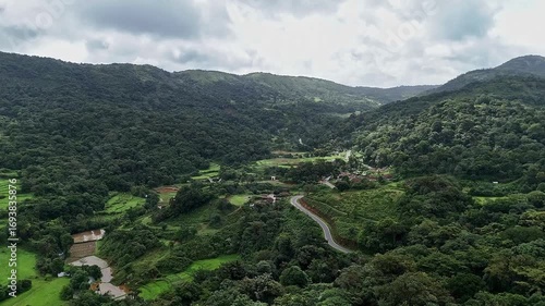 A cinematic drone shot of a small village and farmlands amidst lush mountains under a cloudy sky