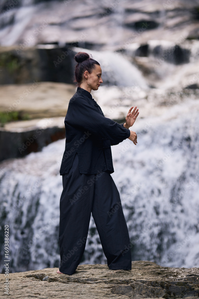Fototapeta premium Woman practicing Tai Chi in nature, wearing a traditional black martial arts uniform, standing gracefully near a waterfall, focused on meditation and balance. Healthy lifestyle in asian culture