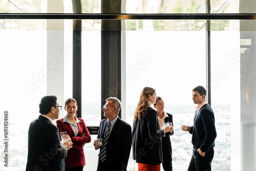Business professionals networking in a modern high-rise with panoramic city views, chatting with drinks during a corporate event by large windows.