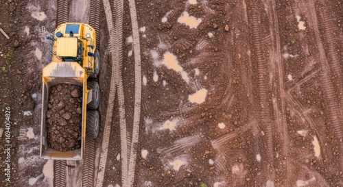 Aerial view of a heavy-duty articulated dump truck filled with earth traversing a muddy construction site with numerous tire tracks and standing water