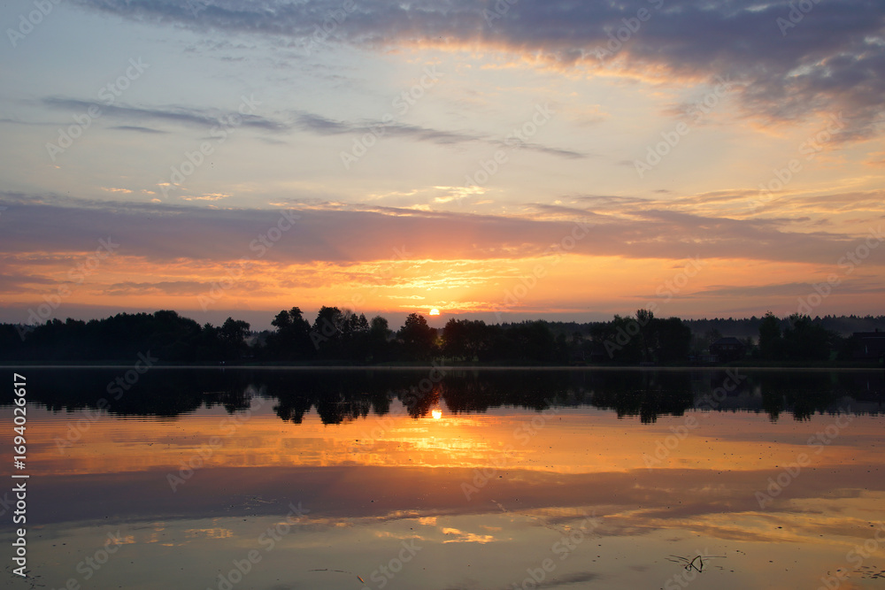 Fototapeta premium Beautiful moments of sunrise. Shot from the surface of the lake, the shore and trees against the beautiful sky. Morning fog. Reflection on the surface of the water.