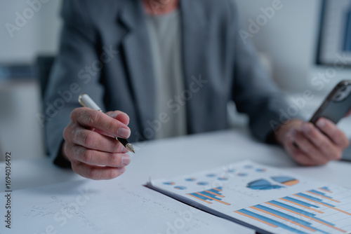 Fotografie Senior businessman holding a pen and using a smartphone, analyzing financial cha