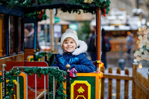 Little preschool girl on a carousel train at Christmas funfair or market, outdoors. Happy child having fun. Traditional xmas market in Germany, Europe. Holiday, children, lifestyle concept