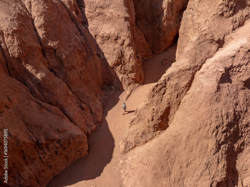 Aerial view of a lone figure walking through the sun-baked, ochre-red canyon of Valle del Arcoiris, the shadows deepening the textures of the arid landscape, San Pedro de Atacama, Chile.