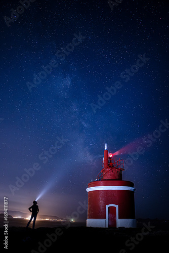 A Night Watcher and the Red Lighthouse Under a Starry Sky. The Milky Way is visible overhead, adding to the cosmic drama. The image captures a moment of human interaction with the immense beauty.