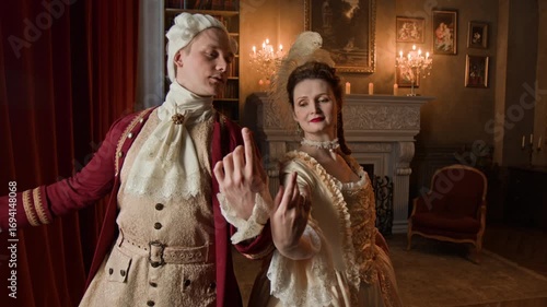 Medium shot of middle-aged Caucasian lady and gentleman in opulent Victorian period costumes performing closing steps of menuet dance then standing still with hands together in 18th century palace
