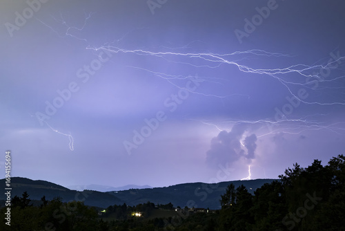 View of jagged lightning bolts electrifying the twilight sky above the dark rolling hills and scattered lights in Graz, Styria, Austria.