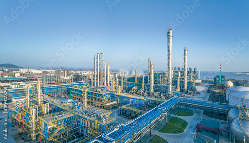 Large modern oil refinery and petrochemical industrial plant with intricate pipelines by the sea under a clear blue sky.