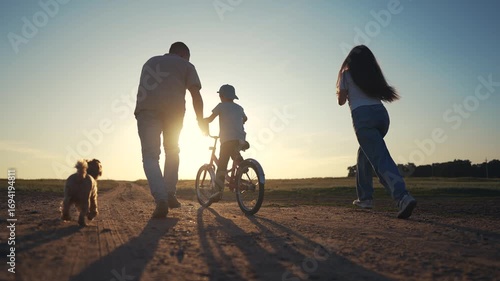 Family on dirt road at sunset. Family teaches their son how to ride bike. A father and child are riding a bicycle. A family on a gravel road lifestyle during sunset.