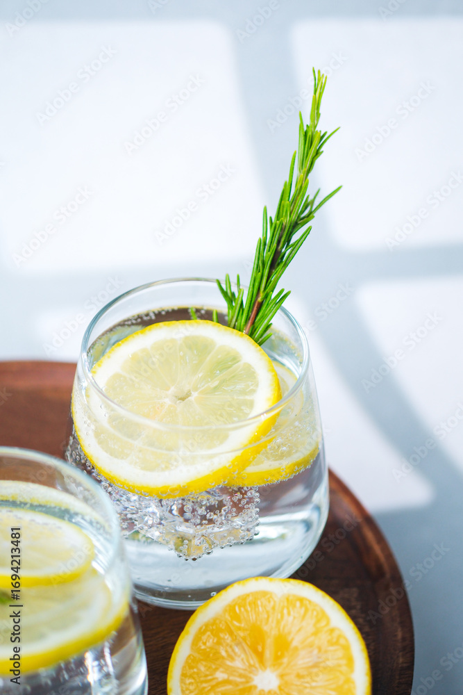 Fototapeta premium Lemon water with rosemary sprigs and ice cubes served in glasses on round wooden table beside fresh citrus halves