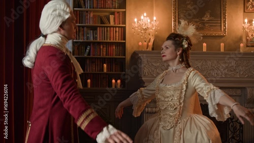 Medium shot of mature Caucasian aristocratic couple in historical costumes performing graceful minuet dance in 18th century royal palace, while attending high society social gathering