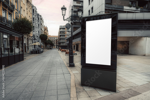 Vertical city billboard mockup with blank white screen on a quiet pedestrian street surrounded by classic and modern buildings, perfect for showcasing urban advertising or branding visuals