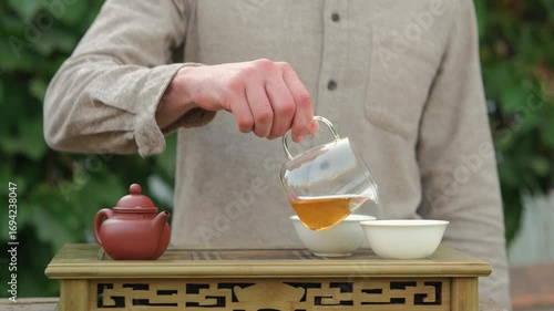 Cropped shot of a man standing behind chaban, pouring tea from glass chahai into two white tea cups, part of an outdoor Chinese tea ceremony