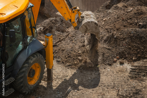 Excavator at construction site performs earthworks dumping soil from bucket on hot sunny day, excavator excavates soil at construction