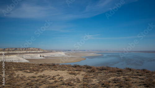The expansive and arid landscape of the Ustyurt Plateau in Kazakhstan, with a large salt flat. The terrain is barren, with low, eroded cliffs visible in the distance under a vast blue sky.