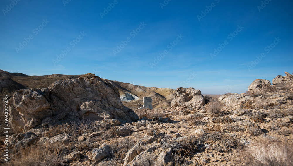 Fototapeta premium A rugged desert landscape stretches across rolling hills, with a striking isolated rock formation standing in the valley below. The arid terrain under a clear blue sky showcases the natural beauty.