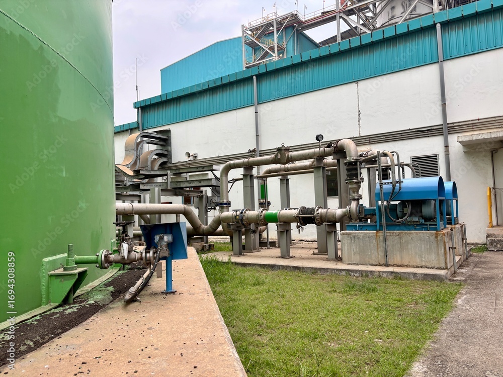 Naklejka premium Industrial landscape featuring a green storage tank and complex piping systems, showcasing infrastructural elements with a backdrop of a large industrial building emphasizing utility and mechanical