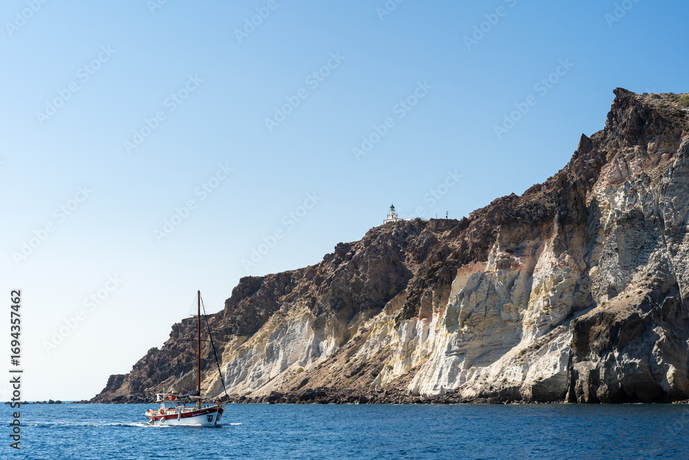 Fototapeta premium Sailboat Cruising by Rugged Volcanic Cliffs with Lighthouse