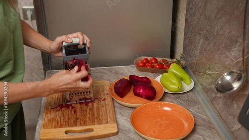 Close-up of a woman grating fresh beets on a box grater over a cutting board, vibrant red shreds falling into place. Preparation of a vegetable dish. Beets for making borscht. Close-up.