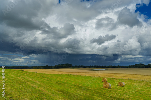 A storm is approaching over the fields, where hay bales are arranged on the fresh green grass. Dark clouds create a dramatic contrast with the peaceful countryside.
