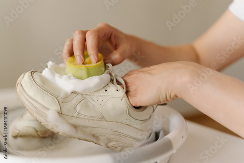 Cropped shot of housewife man cleaning dirty white sneakers with yellow sponge and soap foam in small foldable basin, taking care of footwear hygiene and cleanliness at home in bathroom, close-up.