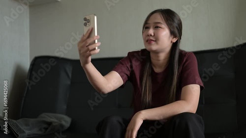 Happy young Asian woman talking on phone and having video call meeting while sitting on sofa at home living room
