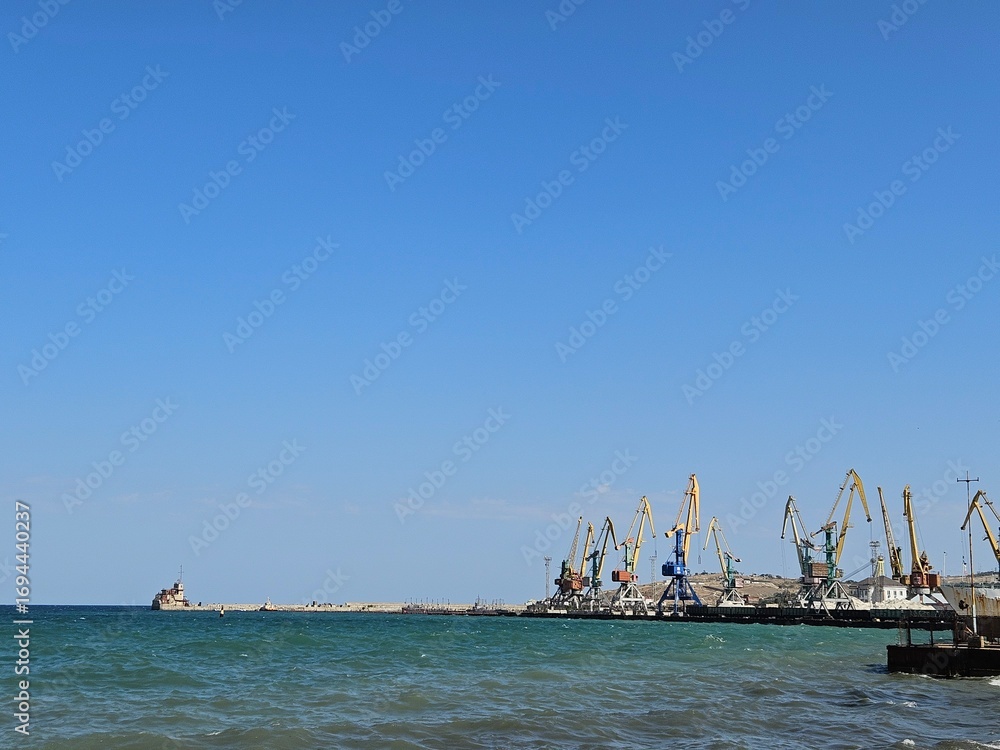 Fototapeta premium Seascape of an industrial port with cargo cranes against a vast blue sky. Perfect for content on global trade, logistics, and the shipping industry. Excellent copy space.