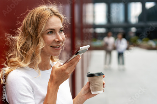 Obraz na plátně Businesswoman using smartphone voice assistant and drinking coffee outdoors