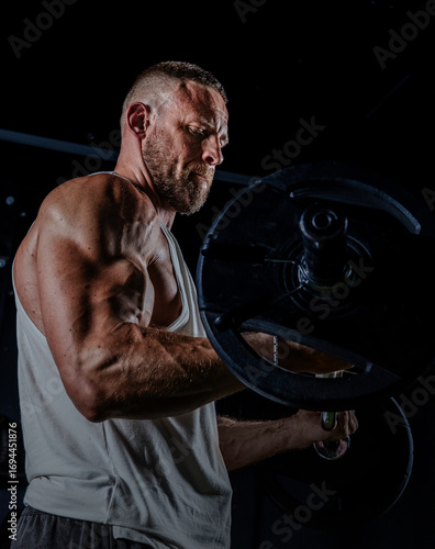 Muscular man training with weights. Brutal strong athletic men pumping up muscles workout in dark gym.