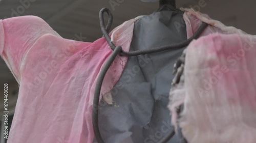 Close-up of a bandaged mummy skull wearing a blue surgical face mask with red stains. Halloween yard decoration with big eyes, tattered gauze, and a suburban house background.