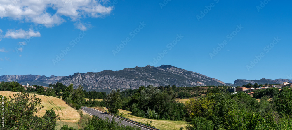 Obraz premium Rugged beauty of the Montsec mountain range in Catalonia, Spain. Scene includes rolling hills, a winding road, and a clear blue sky, creating a picturesque and tranquil landscape typical of the region