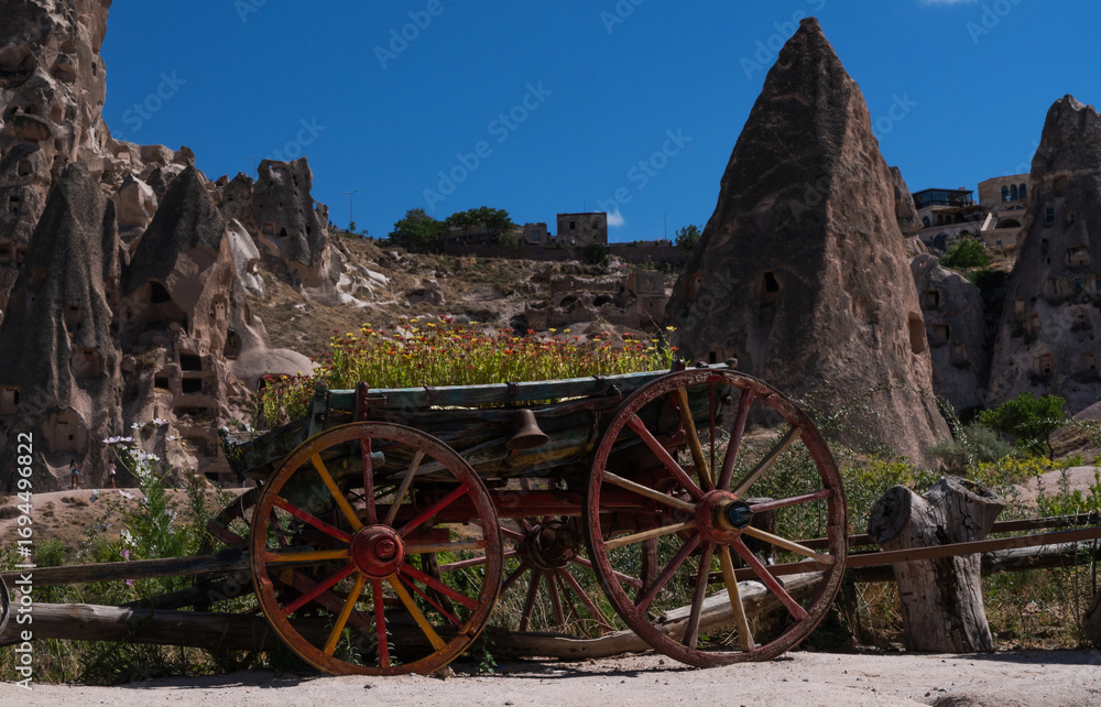 Obraz premium An old wooden cart with vibrant red and yellow wheels stands against a backdrop of unique fairy chimneys and a hill adorned with flowers under the azure sky of Cappadocia.